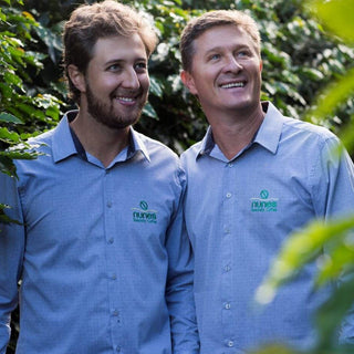 Two men in blue Goshen Coffee shirts smiling among coffee plants on a Brazilian farm