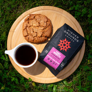 Funky Mamacita coffee bag on a wooden board outdoors with a pastry and a cup of coffee nearby.