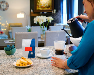 Person in blue pours hot water from a kettle into a cup at a kitchen counter beside an Il Primo coffee bag and cookies