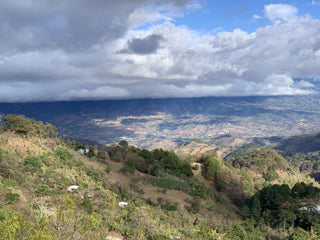 Stormy clouds roll over a mountain valley with distant towns and terraced hills.