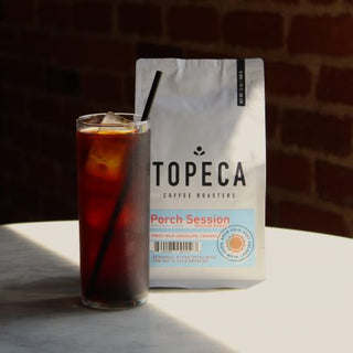 Porch Session Blend bag beside a glass of iced coffee on a white table against a brick wall