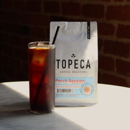 Porch Session Blend bag beside a glass of iced coffee on a white table against a brick wall