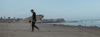 Surfer in a black wetsuit walks barefoot along the beach carrying a white surfboard.
