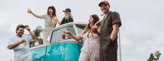 Group of friends celebrating with colorful drinks on a teal vintage van branded Bird Rock Coffee Roasters.