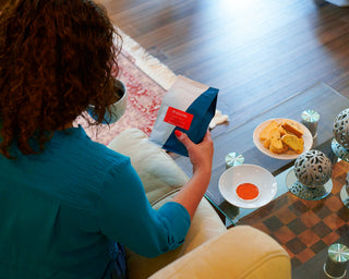 Back view of a person in teal holding a Parisi Italiano coffee bag beside a cup and cookies on a glass table