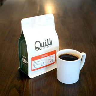 Quills Coffee Inkwell bag on a dark wooden table with a white mug beside it