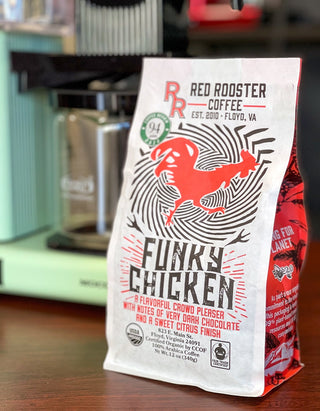 Angled shot of Organic Funky Chicken bag on a wooden counter with roasted coffee beans visible in a cup beside it.
