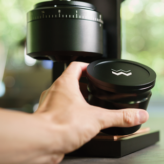 Close up of a hand holding the black lid with a white logo beside The Key compact burr grinder.
