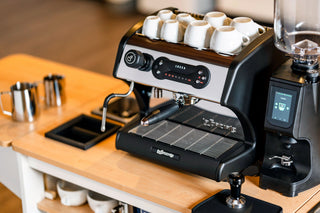 Close-up of a LUCCA espresso machine with white cups on top and a grinder on the counter.