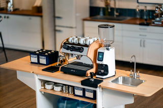 Espresso machine on a kitchen counter with stacked white cups and a coffee setup.