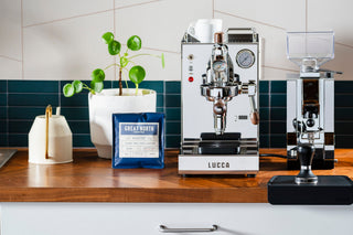 Wide shot of LUCCA Solo espresso machine on a wooden counter with a coffee bag and plant in the background.