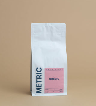 Tall white coffee bag against a warm tan backdrop with a pink SEISMIC label and coordinates.