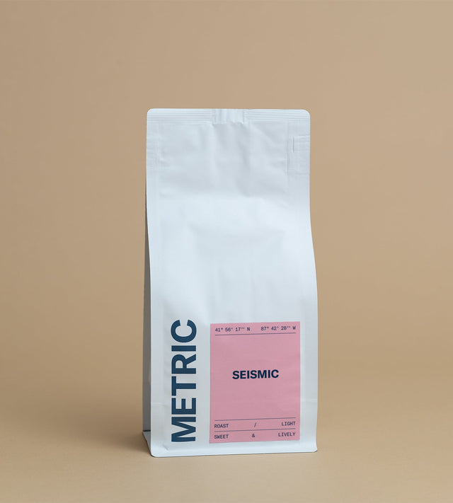 Tall white coffee bag against a warm tan backdrop with a pink SEISMIC label and coordinates.