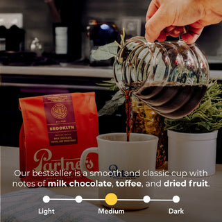 Hand pouring brewed coffee from a glass carafe into a white mug beside a Brooklyn coffee bag