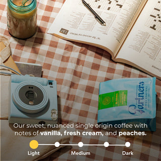 El Ramo coffee bag on a red and white checkered table beside a light blue compact camera and an open crossword book with a pen.