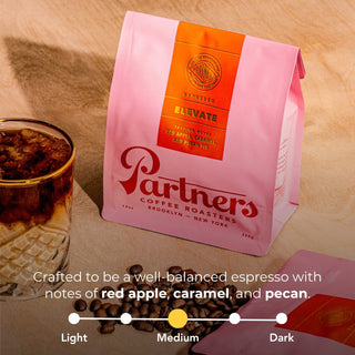 Pink Partners Coffee Elevate espresso bag with an orange label beside a glass of iced coffee and loose beans