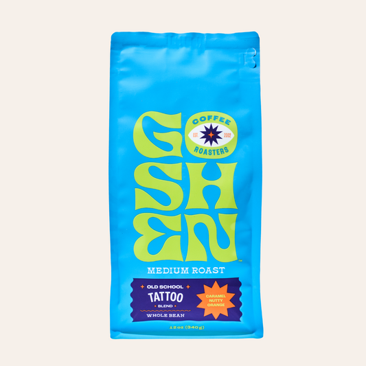 Blue Old School Tattoo whole bean coffee bag with medium roast label beside a cup of coffee on a colorful table