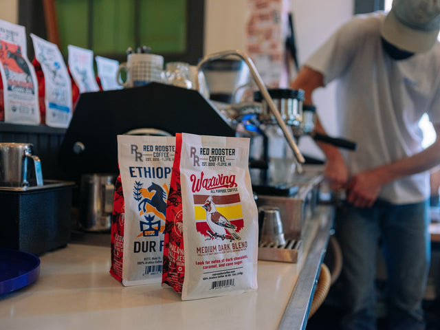 Waxwing All-Purpose Blend bags on a cafe counter with a barista at work in the background.