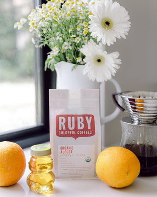 Fresh bouquet of white daisies in a white pitcher sits beside a Ruby Organic August Seasonal Blend bag on a windowsill