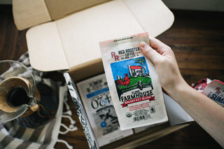 Hand holding Floyd Farmhouse Breakfast Blend bag by Red Rooster Coffee in an open box with other coffee bags.