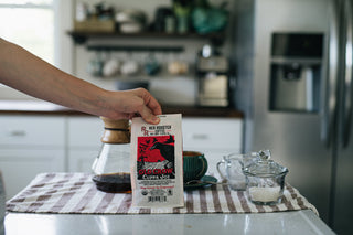 Person holding Old Crow coffee bag on a kitchen counter beside a glass carafe and mug