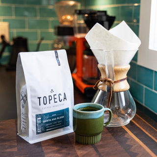 Topeca Santa Ana Blend coffee bag on a wooden counter with a pour over dripper and a teal mug in the foreground.