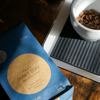 Blue Hunky Dory Organic Decaf bag beside a white cup of beans and a scale on a wooden surface