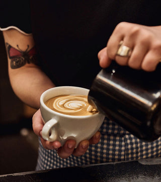 Barista pours milk into a white ceramic cup, creating latte art from an espresso.