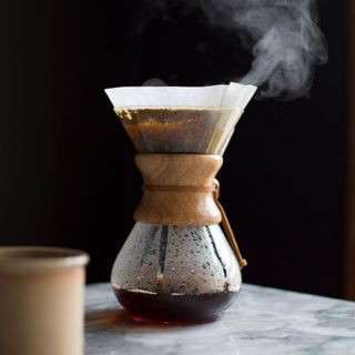 Pour over coffee carafe with a wooden collar and cone filter, steam rising on a marble table