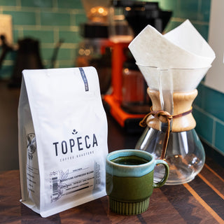 Topeca Signature Espresso Blend bag on a wooden counter beside a glass pour over and a green ceramic mug.