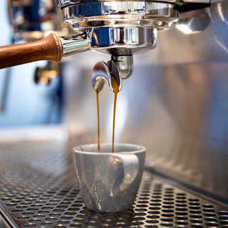 Espresso streams from a portafilter into a white cup with crema forming on a professional espresso machine.