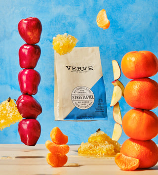 Streetlevel coffee bag amid red apples, orange segments, and honeycombs against a bright blue background