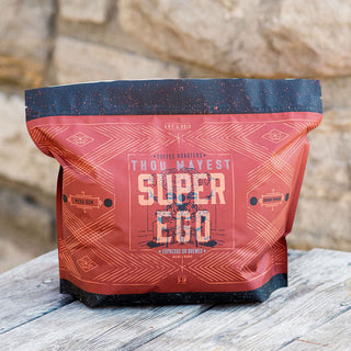 Super Ego coffee bag in red packaging sits on a weathered wooden table against a stone wall.