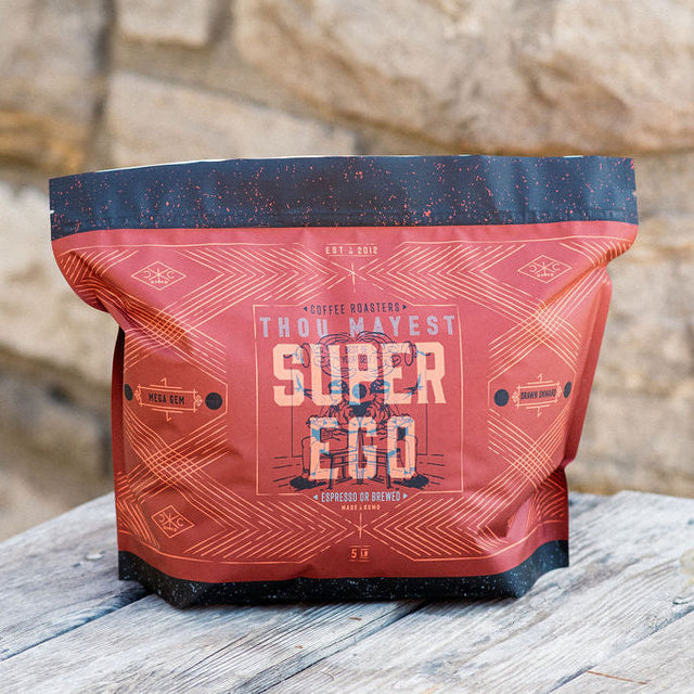 Super Ego coffee bag in red packaging sits on a weathered wooden table against a stone wall.