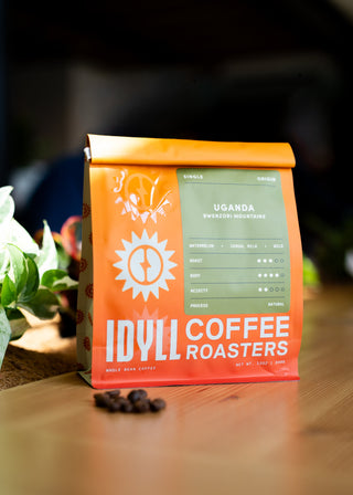 Uganda Rwenzori Natural coffee bag in bright orange with green origin label and sun motif on a wooden table with beans.