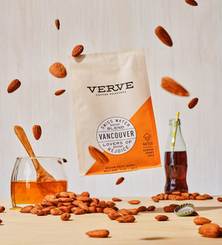 Vancouver Swiss Water Decaf coffee bag on a light wood surface surrounded by floating almonds and a bottle with amber liquid