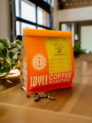 Bright orange Verdancy Spring Blend coffee bag with a yellow label and plant illustration on a wooden surface