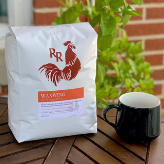 Waxwing All-Purpose Blend bag on a wooden table beside a black mug, with a plant in the background.