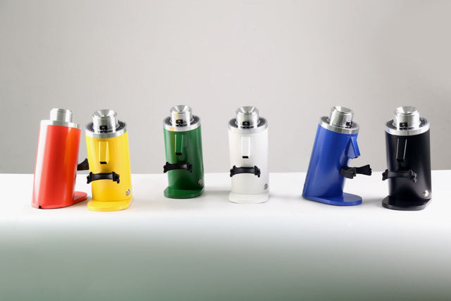 Six colorful Turin DF64 Gen 2.5 single dose grinders lined up on a white table viewed from the side.