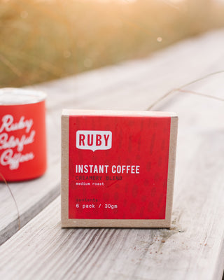 Front view of a Ruby Instant Coffee box beside a red mug on a weathered wooden table.