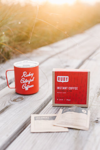 Two Creamery label cards lean on a Ruby Instant Coffee box beside a red mug on a sunlit wooden table.