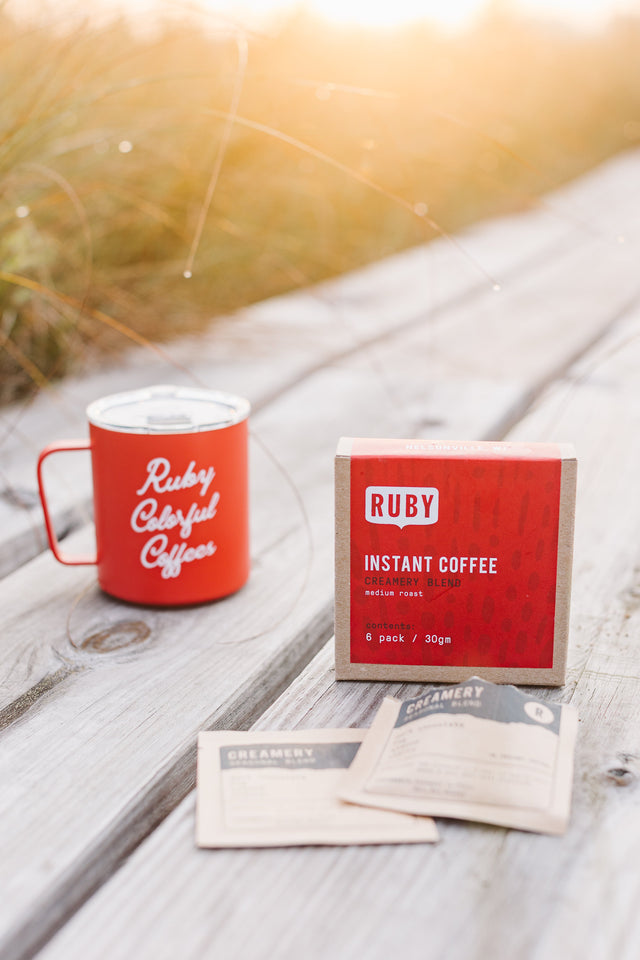 Two Creamery label cards lean on a Ruby Instant Coffee box beside a red mug on a sunlit wooden table.