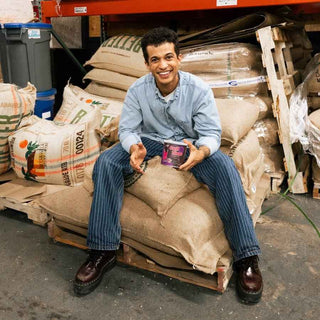 Smiling man in a light denim shirt sits among burlap sacks in a warehouse, holding a Call Time coffee can