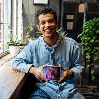 Smiling man in a denim shirt sits by a window, holding a Call Time coffee can toward the camera.