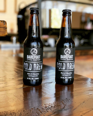 Two Barefoot Cold Brew bottles on a wooden table with a blurred cafe backdrop.