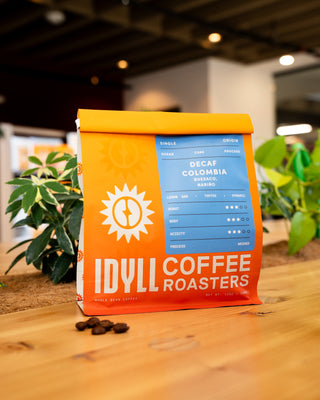 Orange Decaf Colombia Buesaco coffee bag with a blue label on a wooden table beside a leafy plant