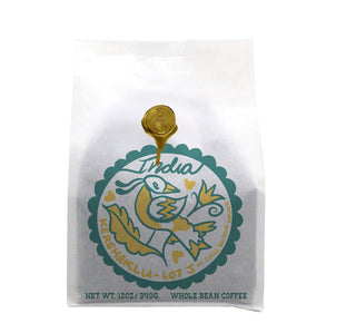 White coffee bag with teal circular logo and a gold seal on top reading India Kerehaklu Coffee Blossom Washed Lot J