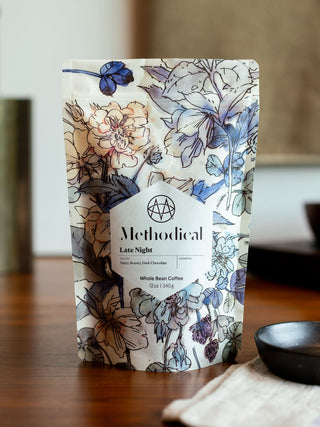 Floral patterned Methodical Late Night coffee bag on a wooden table with a dark bowl nearby