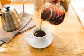 Hand pours dark roast coffee from a glass carafe into a small white cup on a wooden table.