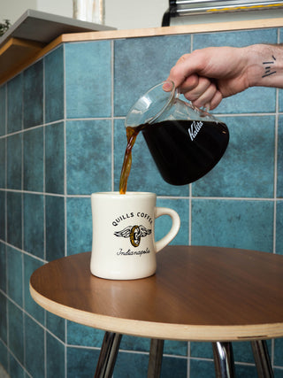 Pour of coffee from a glass Kalita carafe into a Quills Coffee Indianapolis mug on a blue tiled counter.
